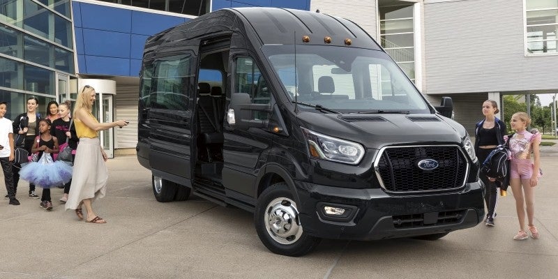 Black Ford Transit van with open door as a woman, children, and dancers gather around it.