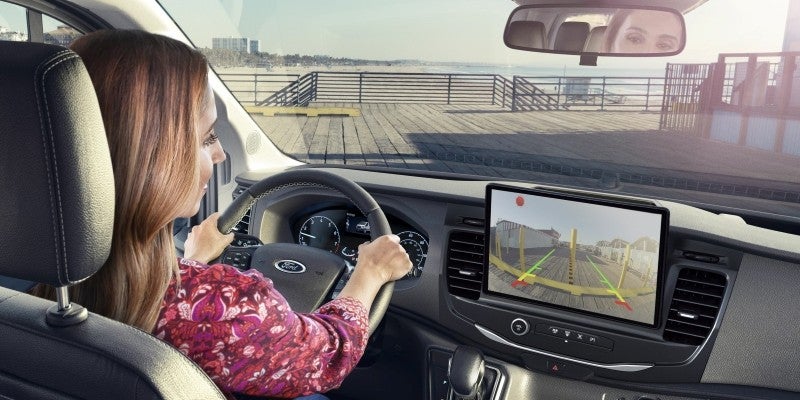 Woman driving a Ford vehicle, using the rearview camera display showing a pier with parking guidelines. Beach and ocean are visible outside.