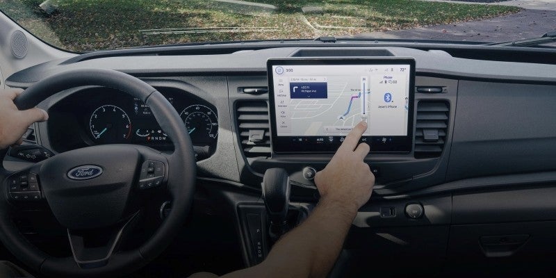 Driver's hands on a Ford steering wheel, interacting with a large touchscreen displaying navigation and phone connection.