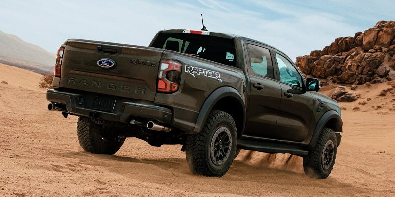 Dark Ford Ranger Raptor pickup truck driving on sandy desert terrain, seen from the rear three-quarter view.