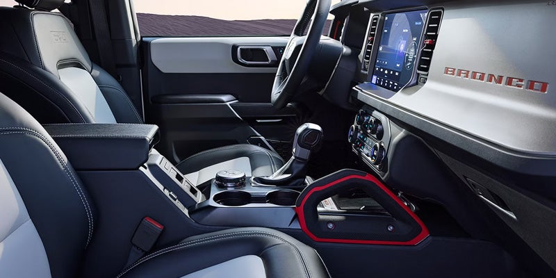 Interior of a Ford Bronco, showing black and white seats, steering wheel, large touchscreen, and red passenger grab handle.