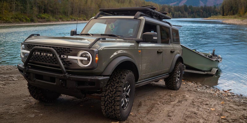 Olive green Ford Bronco with a roof rack parked on a dirt bank next to a river, with a boat partially in the water.