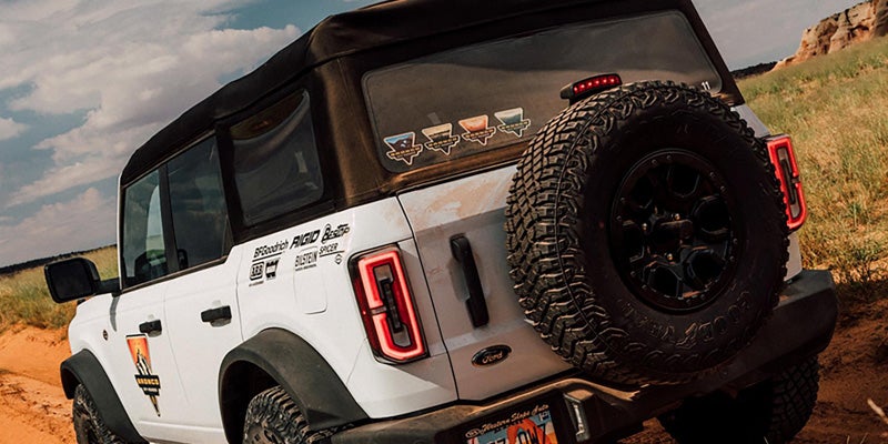 Rear-right view of a white Ford Bronco SUV with a soft top and spare tire, driving on a dirt trail.