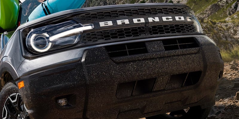 Close-up of a dirty, dark gray Ford Bronco Sport's front end with an illuminated headlight and "BRONCO" on the grille.
