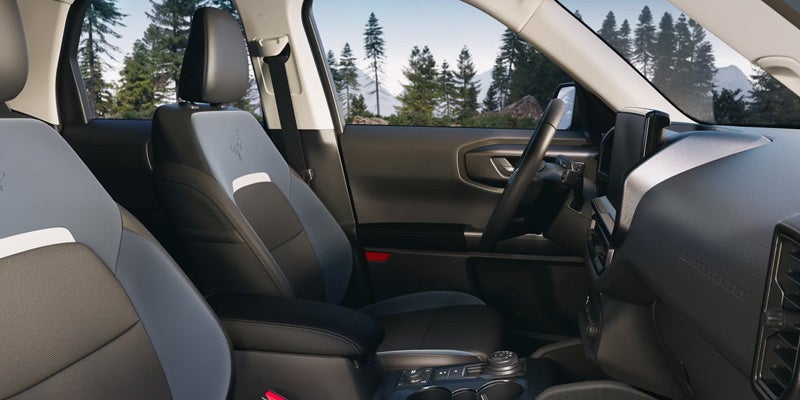 Car interior with two-tone grey and black seats, steering wheel, dashboard, and a view of trees and mountains outside.