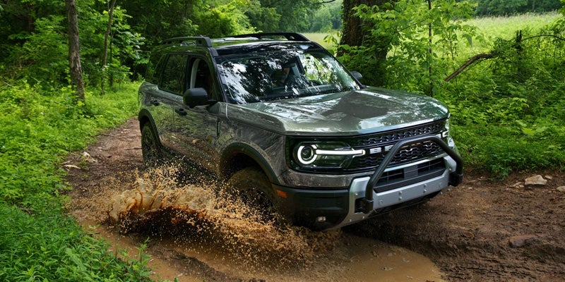 Dark green Ford Bronco Sport SUV drives through a muddy puddle in a forest, splashing mud.