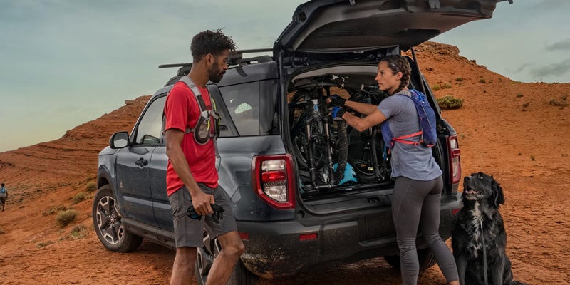 People loading mountain bikes into the back of an SUV in a rocky desert, with a dog nearby.