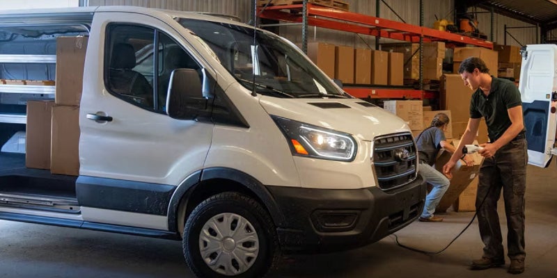 White electric delivery van being charged by a man while another person loads boxes in a warehouse.