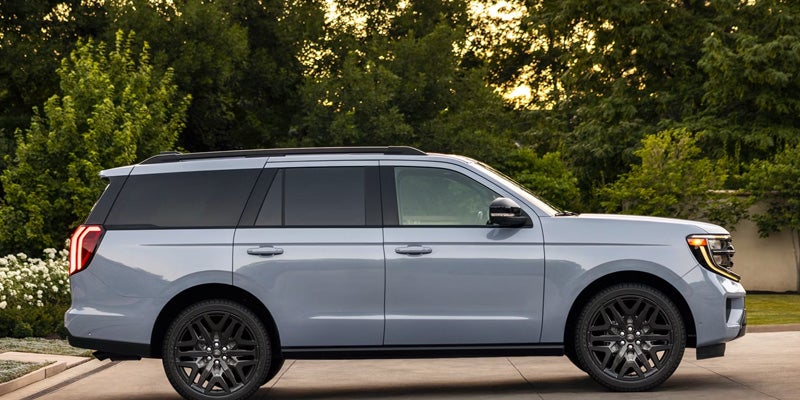 Light gray large SUV shown in profile, parked outside with green trees and golden sunlight in the background.