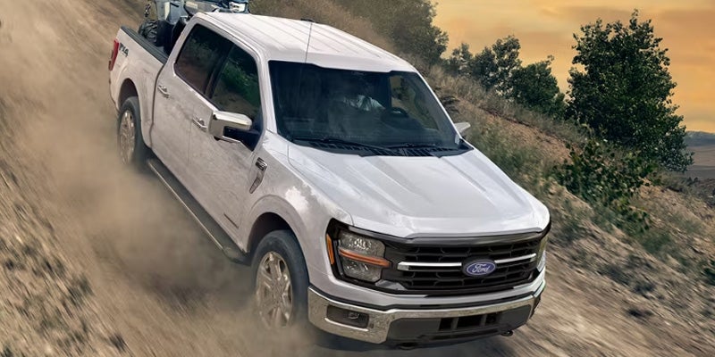 White Ford F-150 pickup truck with an ATV in the bed, driving down a dusty off-road trail at sunset.