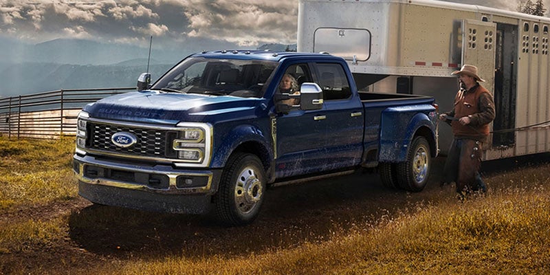 Dark blue Ford Super Duty dually truck towing a livestock trailer, with a driver and a cowboy nearby.