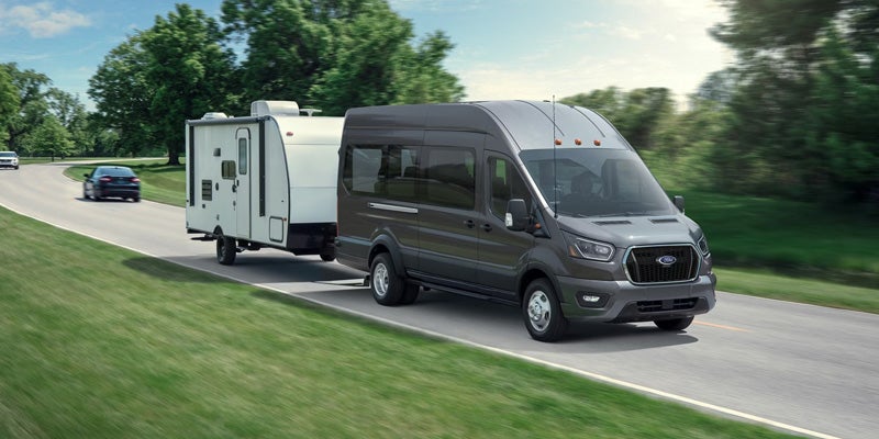 Dark gray Ford Transit van towing a white travel trailer on a winding road with trees.