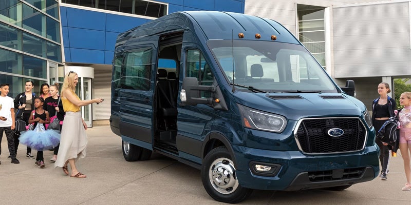 A woman with a remote opens the door of a dark blue Ford Transit van for a group of children and teens.