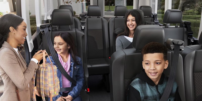 Woman hands a backpack to a smiling child in a passenger van, while two other children sit smiling in the back seats.