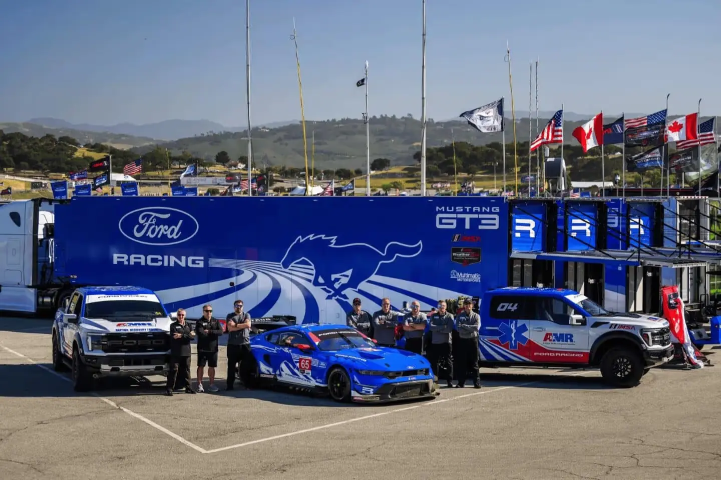 Blue Ford Mustang GT3 race car, two Ford Raptor trucks, and team members in front of a Ford Racing trailer at a track.