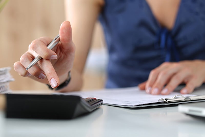 Person's hand holding a pen, pressing a button on a calculator, with paperwork on a clipboard nearby