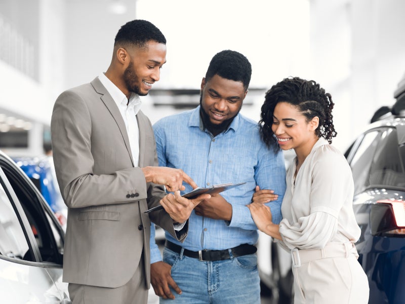 Car salesman points at a clipboard for a smiling couple in a car dealership.