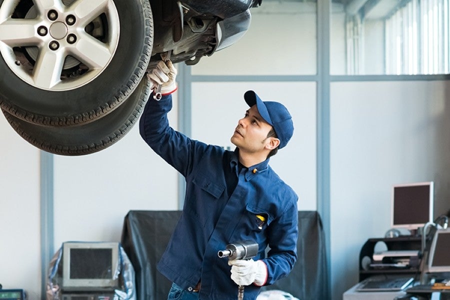 Male mechanic in blue uniform and cap inspecting the underside of a car, holding a wrench and impact gun.